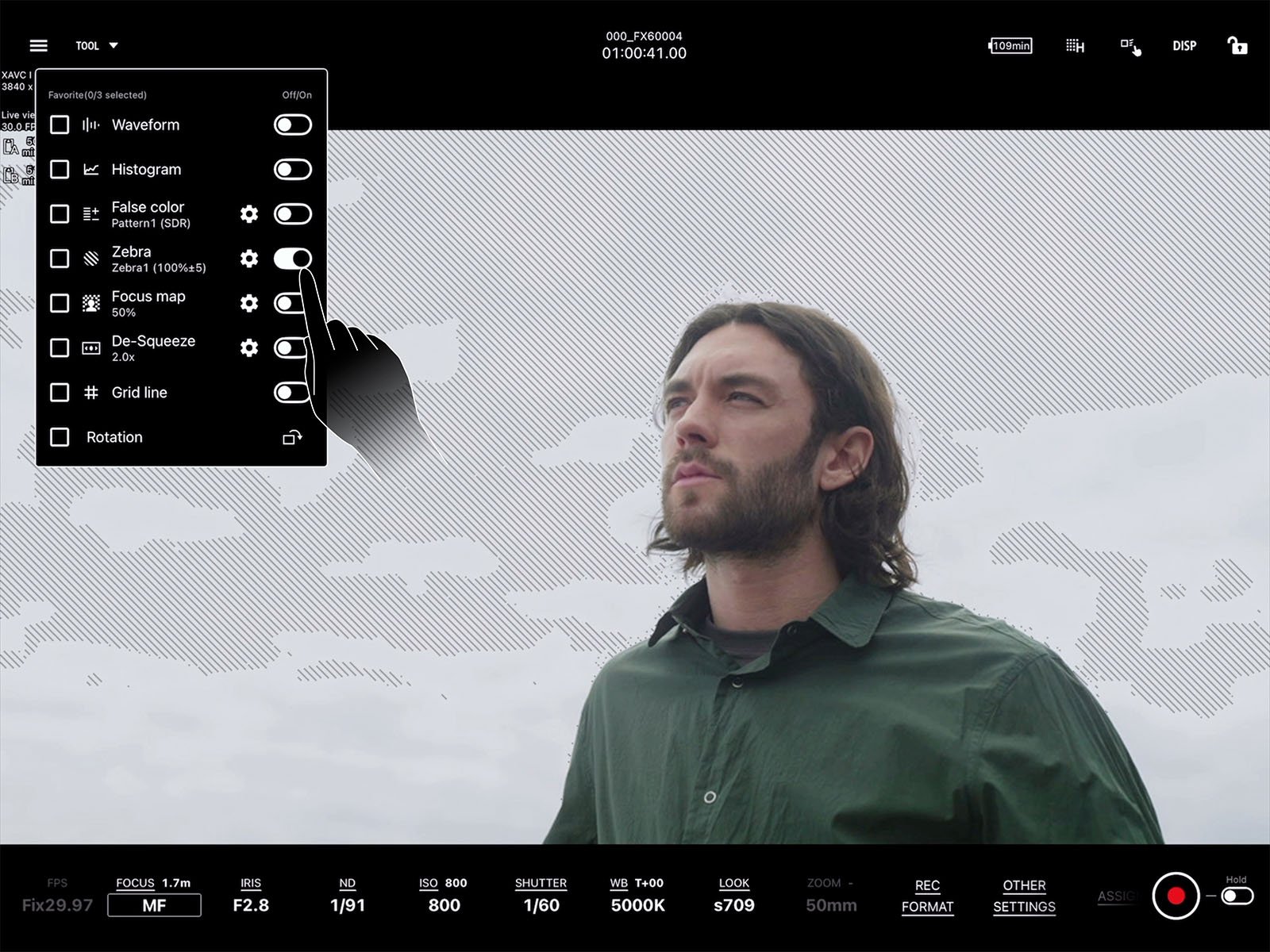 A man with long hair and a beard stands outdoors against a cloudy sky, wearing a green shirt. The image features a film or video camera interface overlay, showing settings such as focus, exposure, and various options for waveform, histogram, and grid.