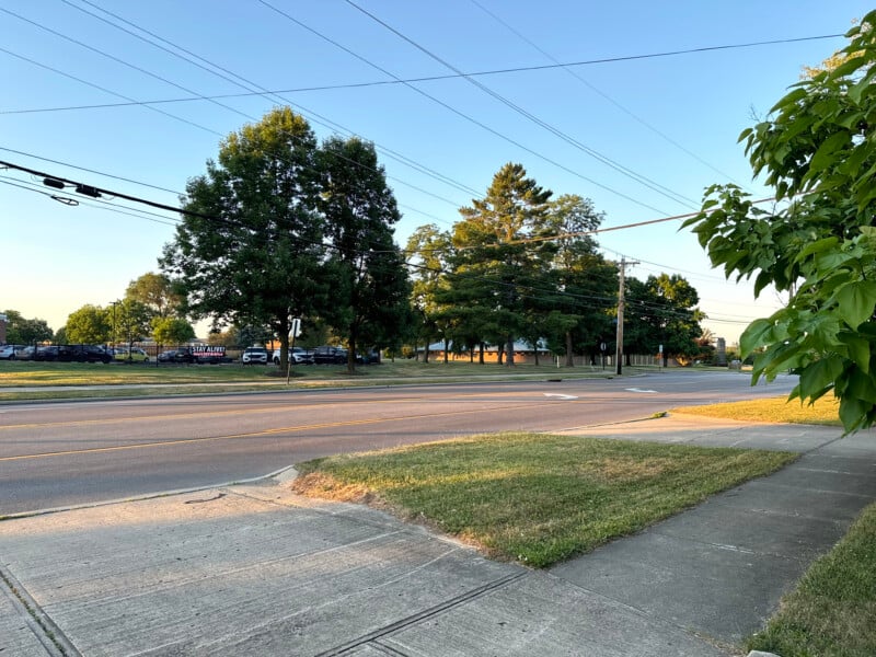 A quiet suburban street with a sidewalk and well-maintained grass. Several large trees line the road, and power lines are visible overhead. It is early evening with clear skies. In the background, a parking lot with vehicles can be seen.