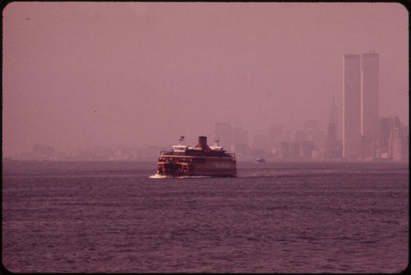 A ferry travels across a body of water toward the city skyline on a hazy day. In the background, the Twin Towers of the World Trade Center stand prominently amidst the dense cluster of buildings, partially obscured by the fog.