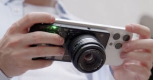 A person holds a smartphone with a detachable camera lens attached to the back, making it look like a digital camera. The smartphone has three camera lenses and is white. The person wears a light-colored shirt. The green indicator light on the attachment is on.