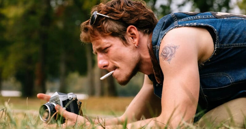 A person with a tattoo on their upper arm is crouched down on grass, focusing on a camera. They have a cigarette in their mouth, sunglasses on their head, and are wearing a sleeveless denim jacket. Trees can be seen in the blurred background.
