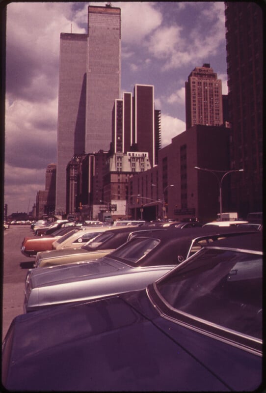 A view of a cityscape with tall skyscrapers under a cloudy sky. In the foreground, a parking lot is filled with various cars, mostly from the 1970s, lined up diagonally. The buildings exhibit a mix of modern and older architectural styles.