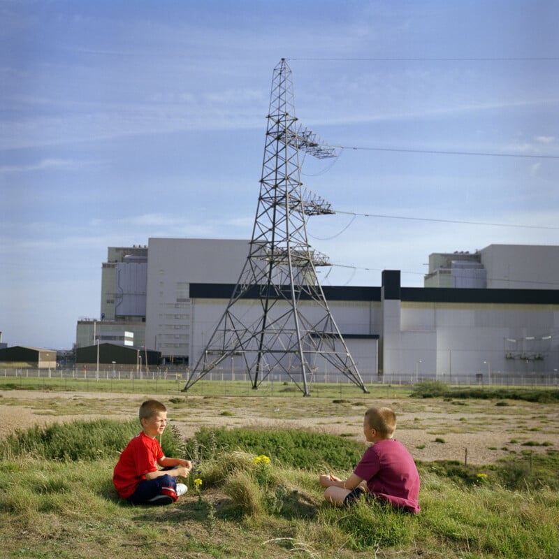 Two children sit on grassy terrain near industrial buildings and a large metal electricity pylon. One child wears a red shirt, and the other wears a purple shirt. The sky is clear and blue, and the overall setting appears expansive and industrial.