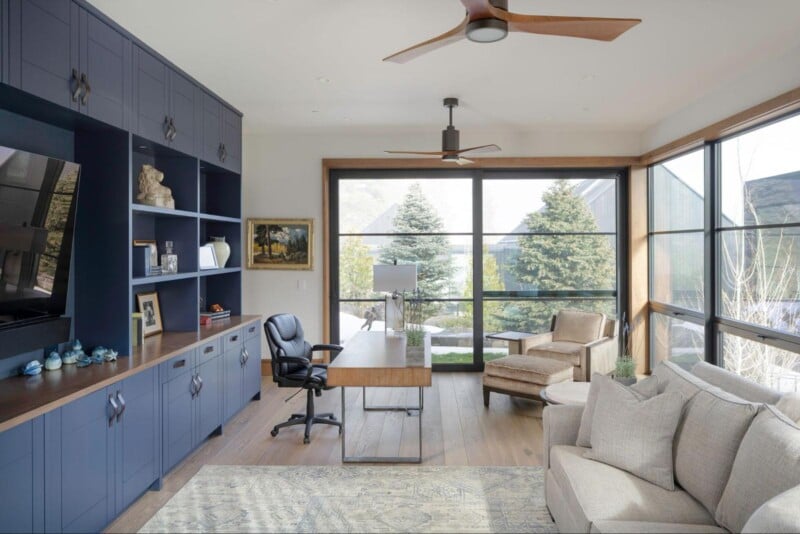 A modern home office with large windows featuring outdoor views, navy blue built-in shelving, a wooden desk with a black chair, a beige armchair with a matching ottoman, a beige sofa, and a light-colored rug. The room has wood flooring and a ceiling fan.