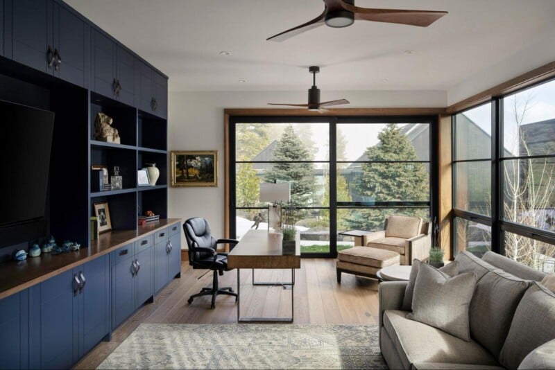 A modern home office with large windows showing a scenic outdoor view. The room has navy blue built-in shelves, a wooden desk, a black office chair, a beige armchair and ottoman, a light gray sofa, and a ceiling fan. Decor includes books, art, and decorative items.