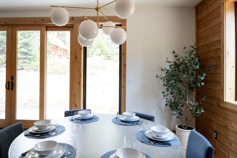 A modern dining area with a round table set for six, featuring white dishes, blue placemats, and a gold chandelier with white globe lights. A potted plant stands in the corner, and large windows showcase a view of trees and a wooden house outside.