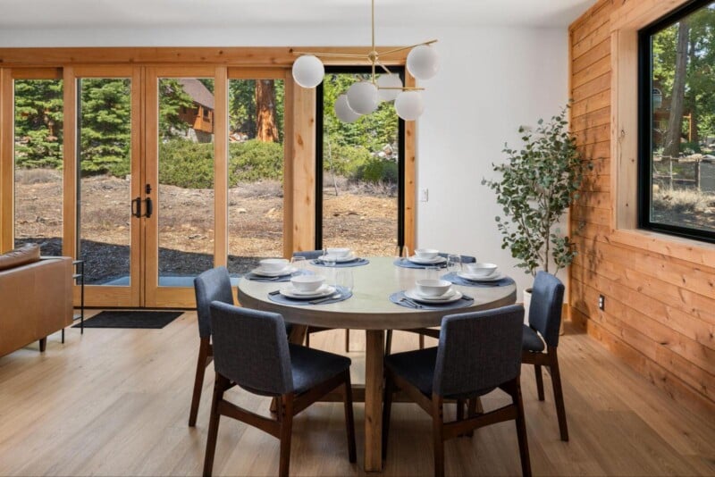 A modern dining area with a round table set for six, featuring white plates, bowls, and cutlery. The room has wooden walls and floor, large glass doors opening to a patio, a potted plant near the window, and a contemporary chandelier above the table.