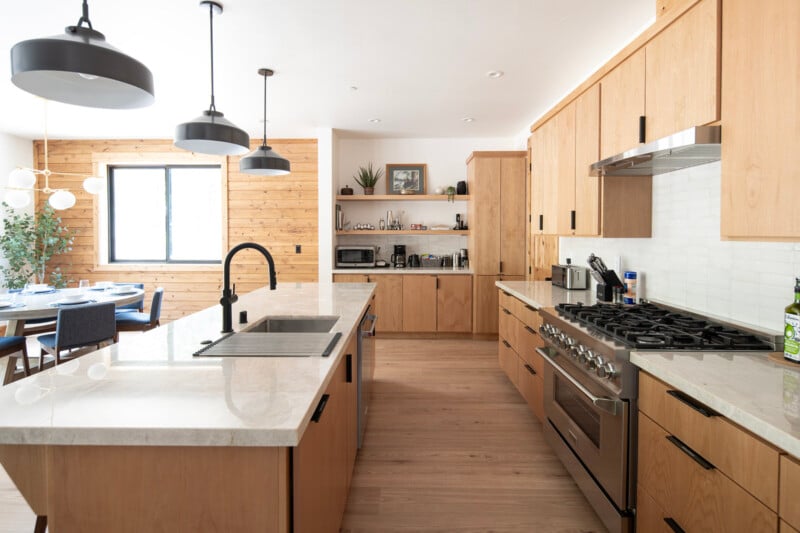 A modern kitchen with light wooden cabinets, white marble countertops, and stainless steel appliances, including a large oven and gas stove. Pendant lights hang from the ceiling. The kitchen leads to a dining area with a wooden wall and a window.