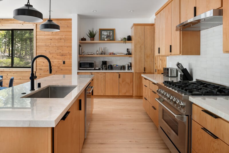 Modern kitchen with light wooden cabinets, stainless steel appliances, and a large island with a black faucet. The space features open shelving with kitchen essentials, a window with a view of greenery, and pendant lights hanging over the island.