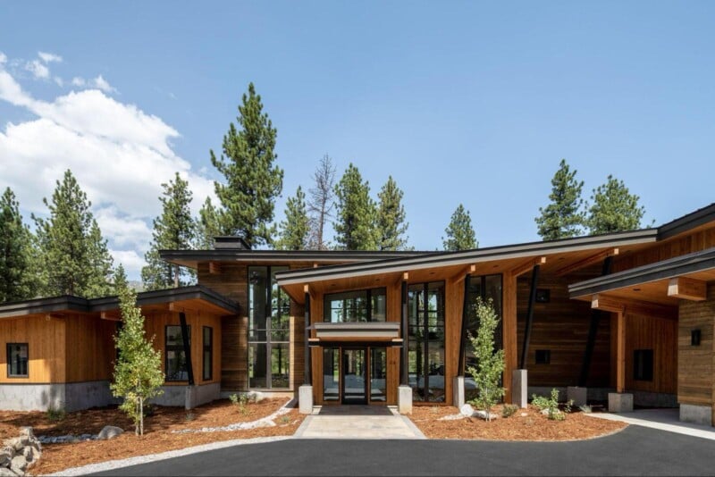 Modern building with large glass windows and wooden exterior set against a backdrop of pine trees and a clear blue sky. The structure features angular rooflines and is surrounded by landscaped areas with small trees and mulch.