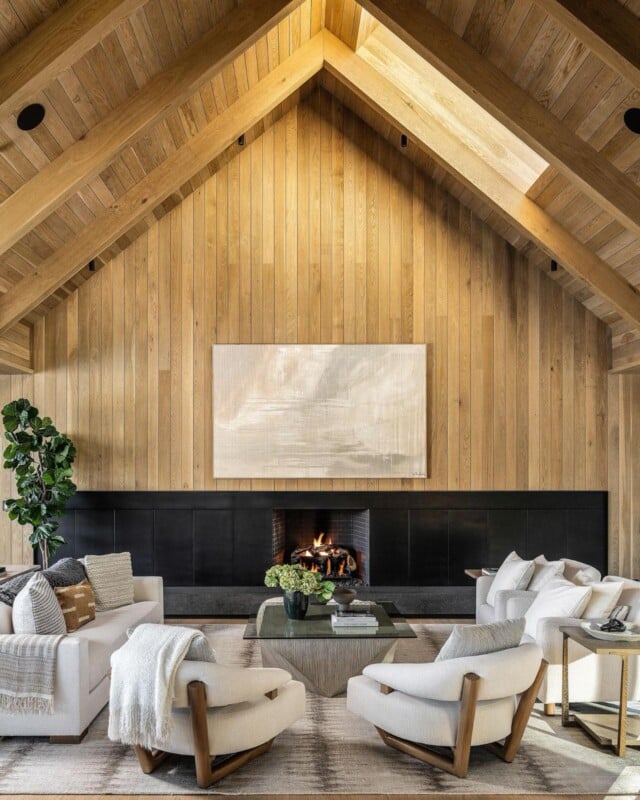 A cozy living room with a high, wooden vaulted ceiling, featuring a central fireplace. The room is furnished with white armchairs and a sofa, arranged around a glass coffee table. Potted plants and a large abstract painting complement the minimalist decor.