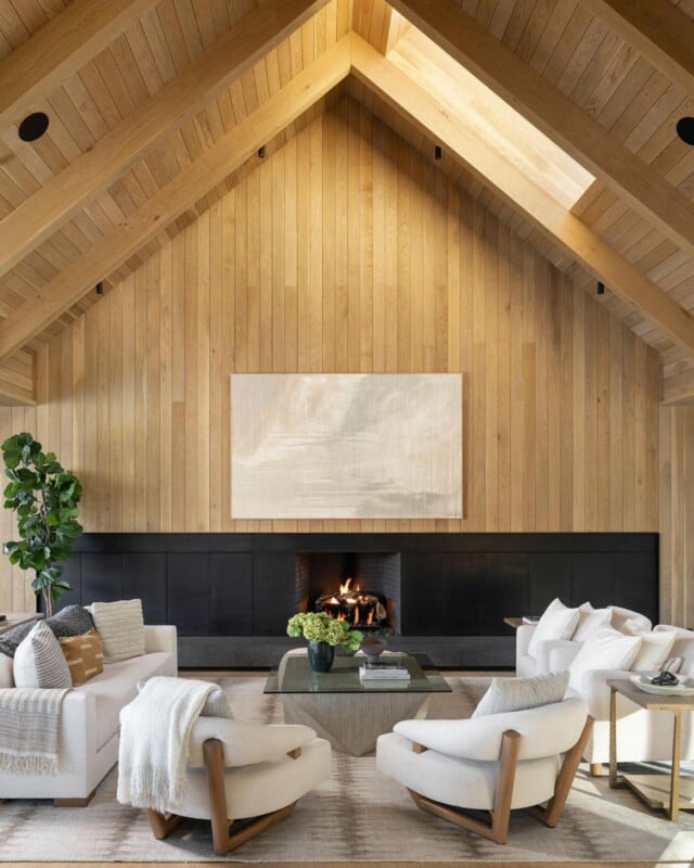 A cozy living room with a high, wooden, vaulted ceiling and a large skylight. The room features a modern fireplace, neutral-toned furniture, including armchairs and a sofa, a glass coffee table, and a minimalist painting on the wooden paneled wall.
