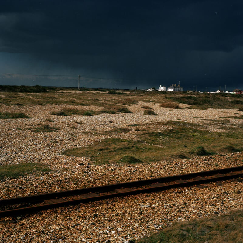 An expansive, gravel-strewn landscape with sparse grass patches is shown under a dark, stormy sky. A rusty railroad track runs horizontally in the foreground. In the distance, a white house with additional buildings can be seen against the dramatic backdrop.