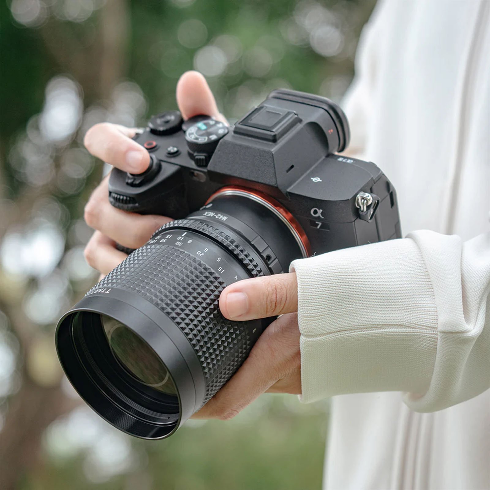 Person holding a digital camera with both hands, wearing a white long-sleeved top. The camera has a large attached lens, and the background is slightly blurred, showing some greenery.