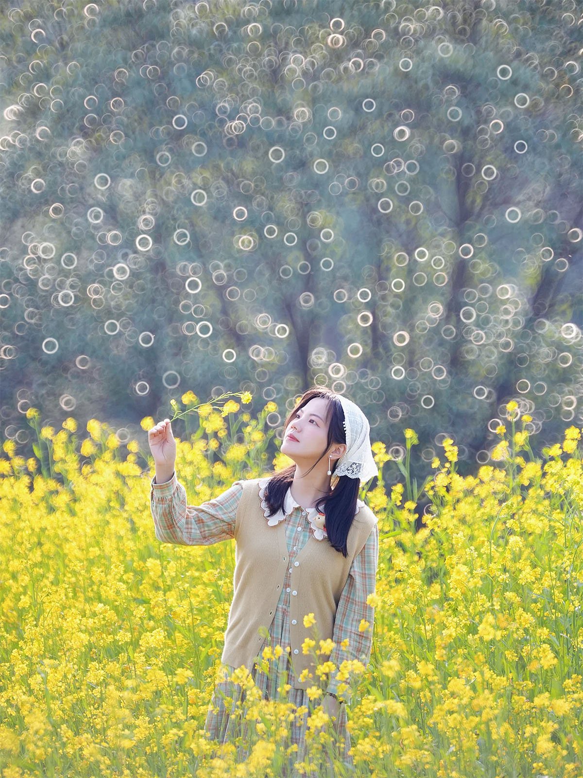 A woman stands in a field of vibrant yellow flowers, holding one close to her face. She wears a light-colored headscarf and a beige vest over a plaid shirt. The background features a dreamy, bokeh effect with circular light patterns and blurry trees.