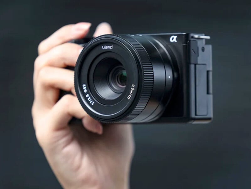 A person holds a black mirrorless camera with a Ulanzi 23mm f/1.4 lens attached. The camera is positioned against a plain dark background. The person's fingers are wrapped around the camera, ready to take a photo.