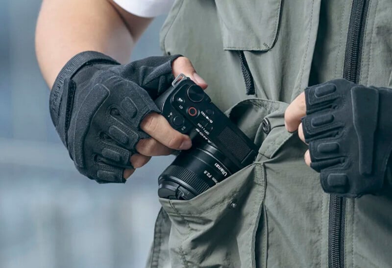 A person wearing a green vest and black tactical gloves is holding a black camera, placing or pulling it out of a pocket on the vest. The focus is on the hands, gloves, and camera, with a blurred background.