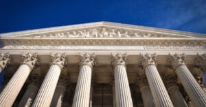 The image shows the facade of the U.S. Supreme Court building, featuring six large Corinthian columns supporting a triangular pediment. Engraved above the columns is the phrase "EQUAL JUSTICE UNDER LAW." Figures are sculpted in the pediment, set against a clear blue sky.