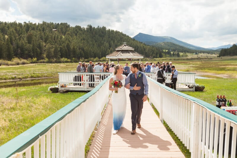A bride in a white dress and a groom in a vest walk hand in hand across a wooden bridge after their outdoor wedding ceremony. Guests gather in the background near a gazebo. The setting is scenic with greenery, a mountain range, and a partly cloudy sky.