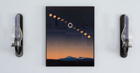 A framed illustration showing the phases of a solar eclipse. The sequence of partial to total eclipse is displayed above a landscape featuring mountain peaks at dusk. The frame is flanked by two wall-mounted oil lamps with glass chimneys.