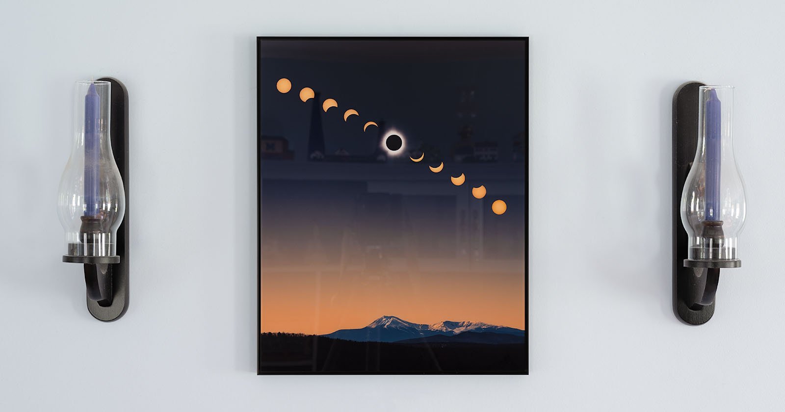 A framed illustration showing the phases of a solar eclipse. The sequence of partial to total eclipse is displayed above a landscape featuring mountain peaks at dusk. The frame is flanked by two wall-mounted oil lamps with glass chimneys.