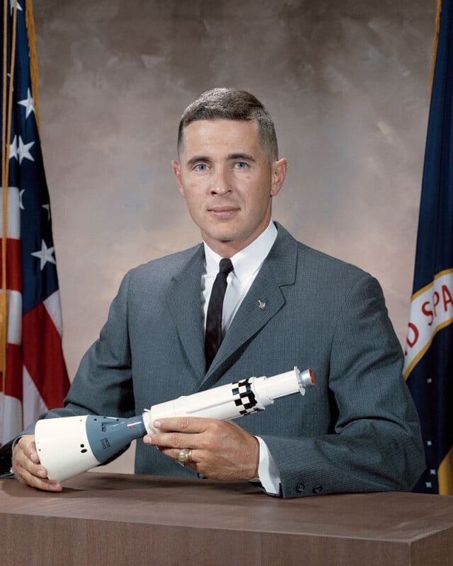 Een man in een grijs pak en zwarte stropdas houdt een modelraket vast terwijl hij poseert voor een officiële foto.  Achter hem staan ​​de Amerikaanse vlag en de NASA-vlag.  De tafel voor hem is leeg, op een modelraket na.