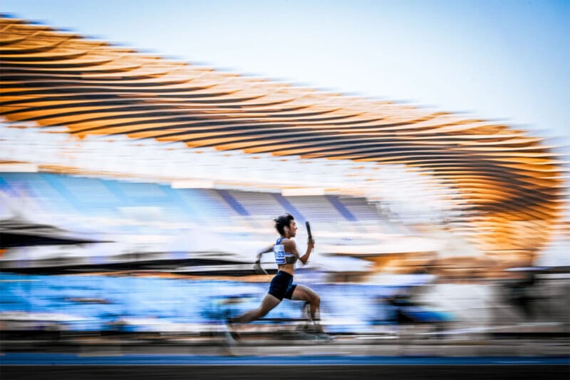 A sprinter in athletic wear is captured mid-run on a track, with blurred stands and a wavy-roofed stadium in the background, conveying a sense of speed and motion.