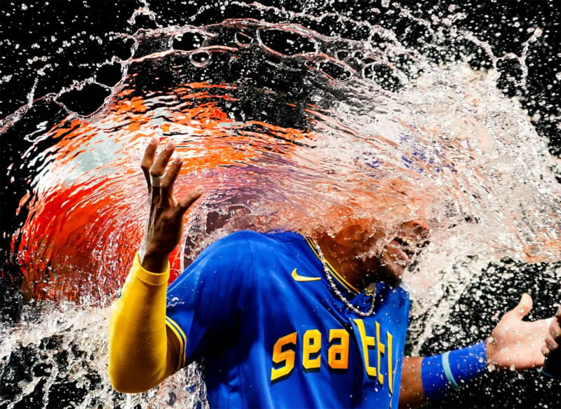 A baseball player wearing a blue "Seattle" jersey and yellow wristbands is hit with a large splash of water, causing water droplets to cascade dramatically around him. The water is vividly illuminated, creating a striking visual effect against the dark background.