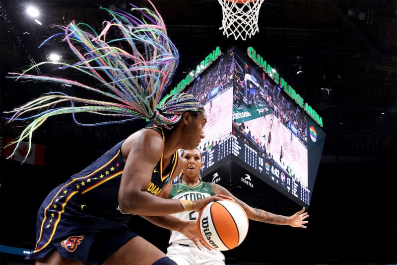 A basketball player with colorful braided hair drives toward the basket in a game. An opposing player defends, and a large scoreboard in the background displays the game in progress at Climate Pledge Arena.
