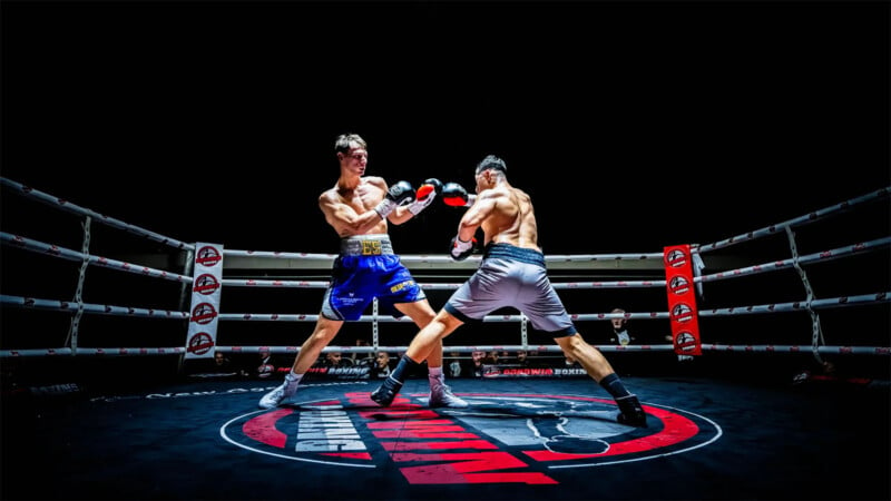 Two boxers are engaged in a match inside a brightly lit boxing ring. One boxer in blue shorts throws a punch, while the other in gray shorts dodges. The ring features a distinctive logo on the floor, and red corner padding is visible in the background.