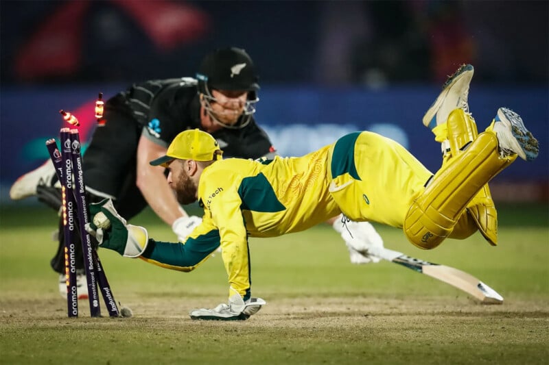 A cricket player in a yellow uniform dives to break the stumps with the ball as a player in a black uniform slides his bat towards the crease to avoid being run out. The stumps are dislodged, and bails are in mid-air. The scene is intense and action-packed.