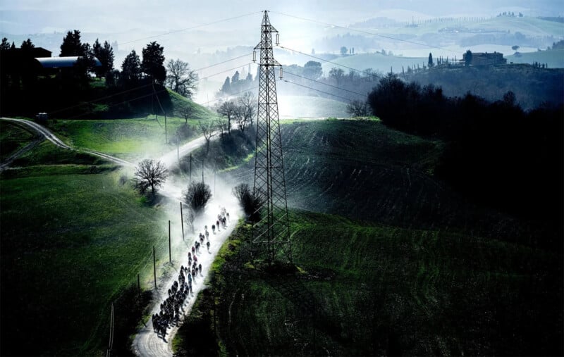 A group of cyclists rides down a winding country road through a lush, green landscape. Morning mist rises from the ground, and an electric transmission tower stands tall beside the road. Rolling hills and scattered trees create a serene background.