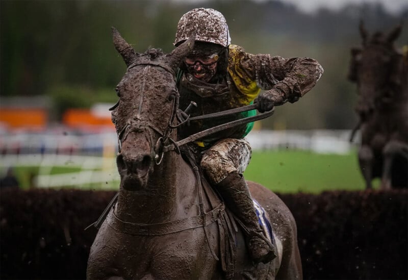 A jockey and their horse covered in mud race toward the camera. The jockey's face is almost unrecognizable due to the mud, and the horse is straining forward with its ears back. The background shows a blurred racetrack with another horse and jockey barely visible.