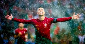 A soccer player in a red and green uniform with the number 7 and a captain's armband celebrates with arms outstretched in the rain. The background shows blurred figures and rain falling heavily, creating a dramatic, triumphant scene.