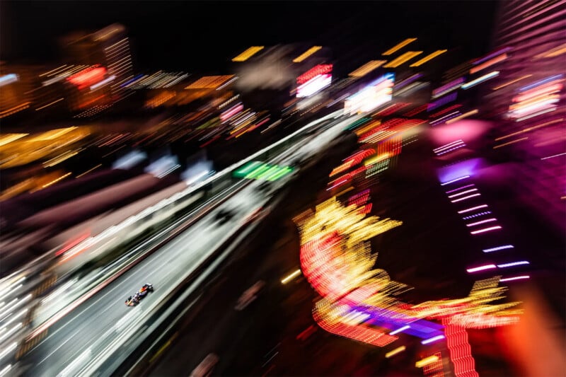 An aerial shot of a nighttime car race featuring a single race car speeding along a well-lit urban track. The background showcases colorful, blurred neon lights from buildings and decorations, creating a sense of motion and excitement.