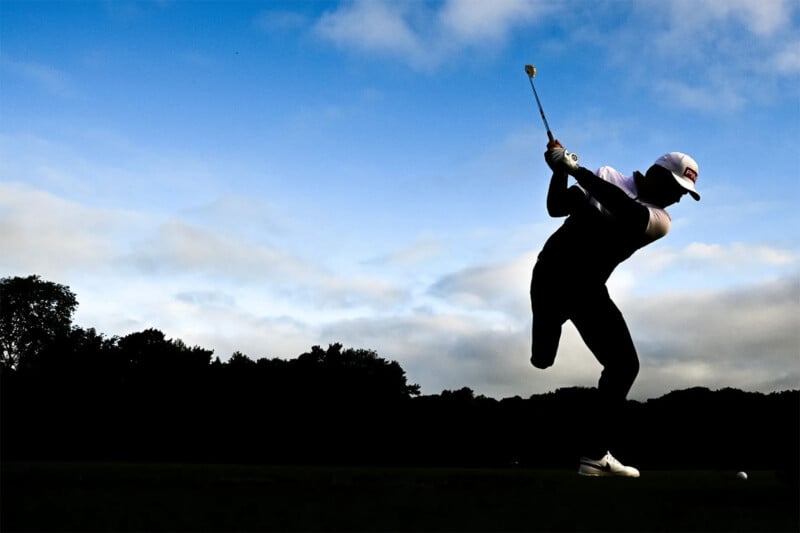 A silhouetted golfer swings a club against a vibrant blue sky with scattered white clouds. The golfer is in mid-motion, emphasizing the dynamic nature of the swing. Trees line the horizon in the background, framing the scene.