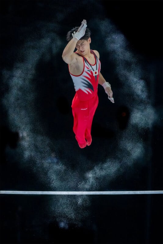 A gymnast in mid-air performs a maneuver on the horizontal bar. Wearing a red and white outfit, they have chalk dust surrounding them, creating a dynamic effect against the black background.
