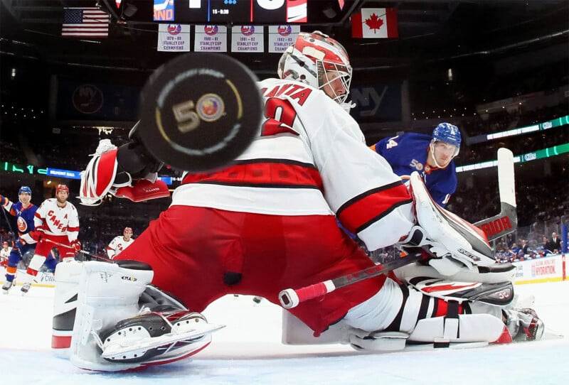 A hockey goalie in red and white gear blocks a shot during a game. The puck, marked with "50", is captured in mid-air near the center of the image. Opposing team players in blue jerseys can be seen in the background on the ice rink.
