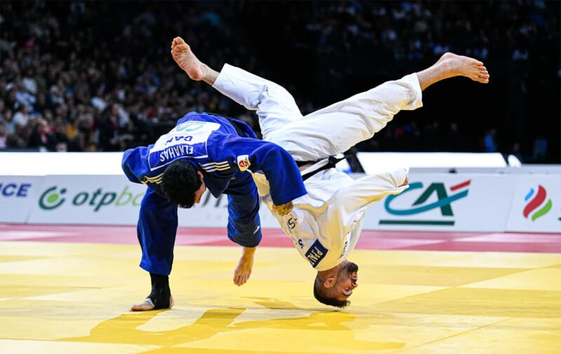 Two judokas are mid-throw during a judo match on a yellow mat in front of a crowd. One judoka in a blue gi is being flipped upside down by their opponent in a white gi. The background shows a large audience and some sponsor logos.