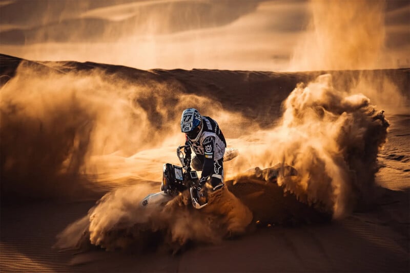 A motocross rider, wearing black and white gear and a helmet, kicks up a massive cloud of sand while riding through a desert. The background features rolling sand dunes bathed in warm, golden light, highlighting the dynamic action in the scene.