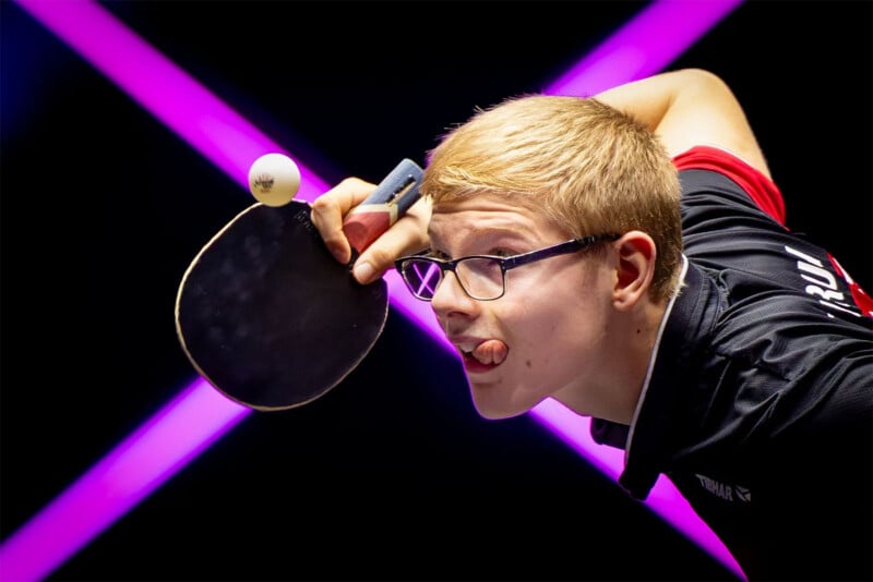 A person with short blonde hair and glasses focuses intently while playing table tennis, tongue out in concentration. They are holding a paddle near their face, preparing to hit the ball. The background features a bright pink "X" shape.