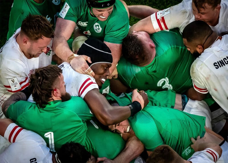 Rugby players from two teams in green and white jerseys are engaged in a scrum during a match. The players' faces show intense concentration and physical effort as they push against each other, creating a tight, bustling formation.