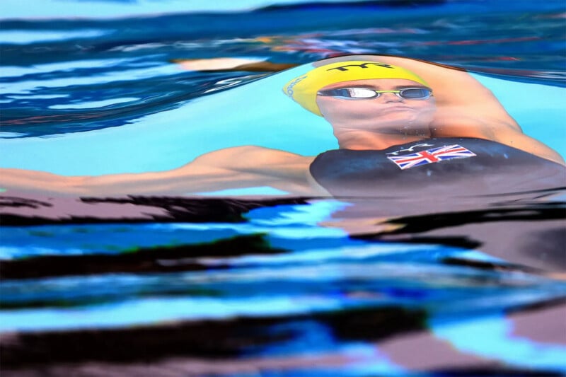 A swimmer wearing a yellow swim cap and goggles performs a backstroke in a pool. The swimmer's right arm is extended while the left arm is by their side. The swimmer's dark swimsuit features the flag of the United Kingdom. The water creates a reflective, wavy effect.