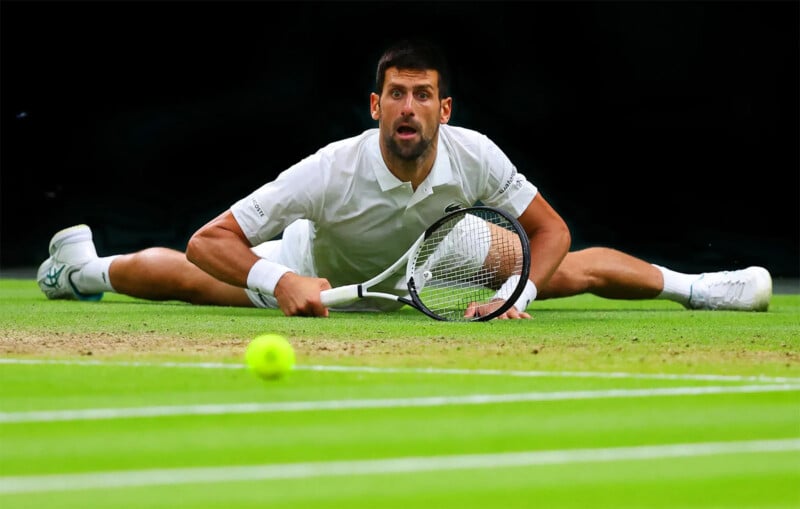 A tennis player in a white outfit is performing a split on a grass court, reaching out with his racket near the ground. A tennis ball is visible in the foreground. The player's expression is intense and focused.