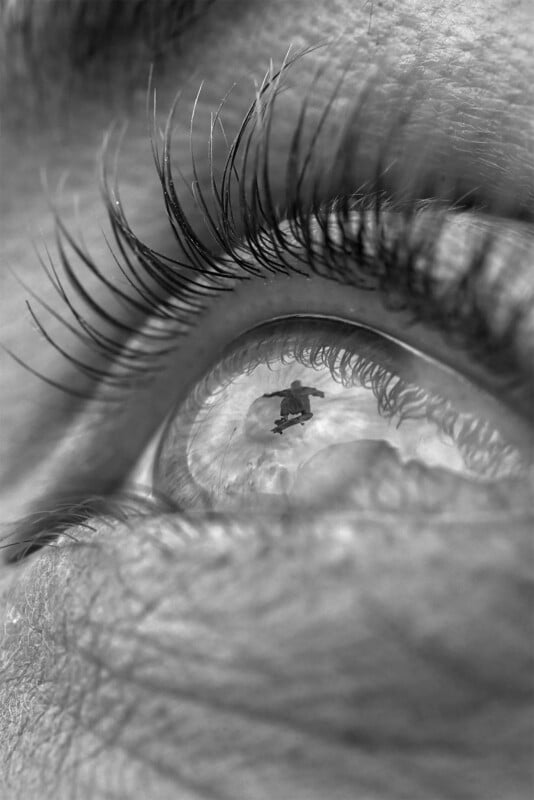 Close-up grayscale image of an eye with long eyelashes. The eye reflects a scene of a person with an umbrella, standing on a surface with clouds in the sky, blending the external view with the inner texture of the eye.