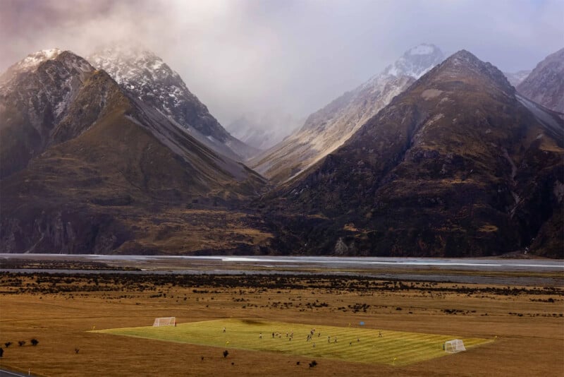 A soccer match is taking place on a grassy field set in a vast, open plain surrounded by towering, rugged mountains with patches of snow near their peaks. The sky above is cloudy, casting a dramatic light over the scenic landscape.