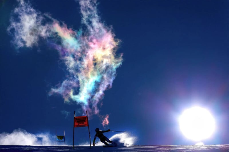 A skier competes in a race under a clear blue sky. The skier, leaning into a turn, leaves behind a colorful spray of snow particles illuminated by the bright sun. Vibrant hues of red, orange, yellow, green, and blue form a striking plume in the air.