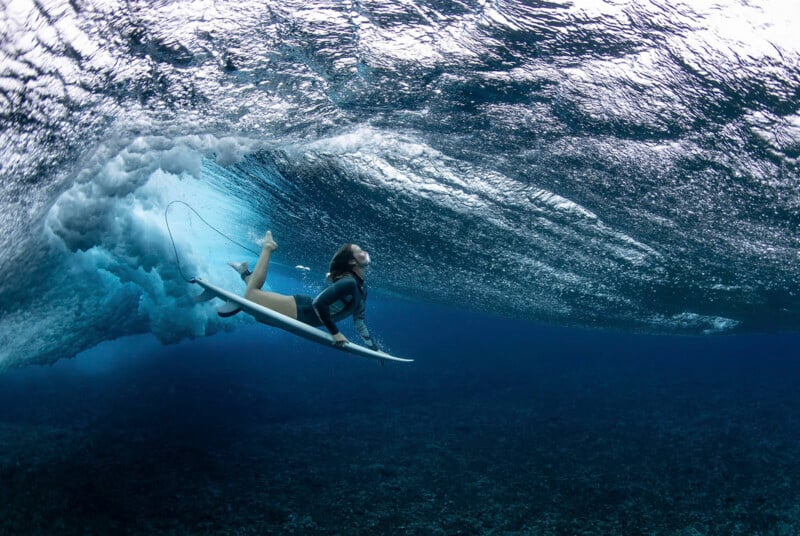 A surfer in a wetsuit rides underneath a crashing ocean wave. The underwater view captures the surfer hanging onto their surfboard, surrounded by swirling bubbles and the deep blue sea. The scene exudes the powerful and dynamic nature of surfing.