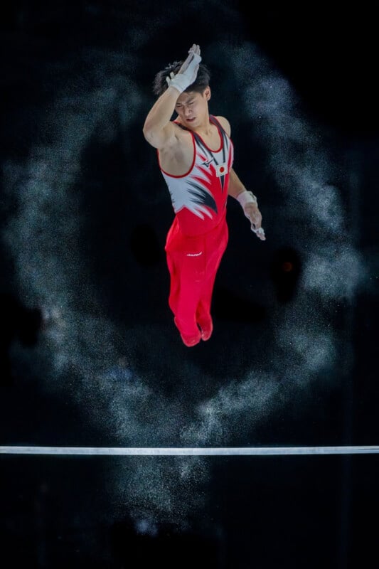 A male gymnast is captured mid-air above a horizontal bar, wearing red pants and a white and red tank top. Chalk dust surrounds him, creating a circular pattern. The background is dark, emphasizing the gymnast and the chalk.