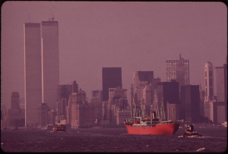 A large red cargo ship sails in the foreground, while the iconic skyline of New York City, including the Twin Towers of the World Trade Center, stands prominently against the hazy, dusky sky in the background. Other boats are visible on the water.